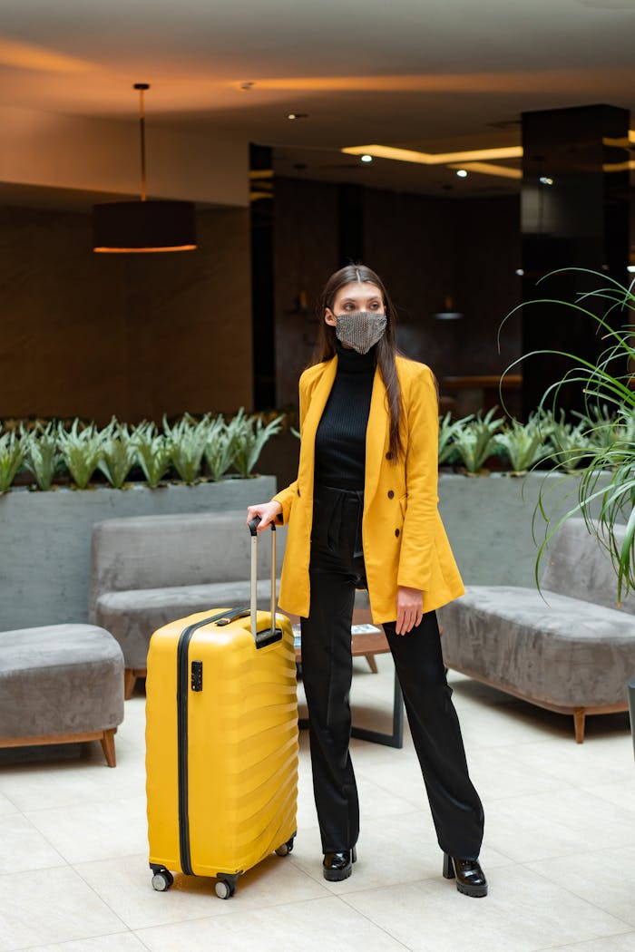 Stylish woman with yellow suitcase and face mask in a modern hotel lobby.